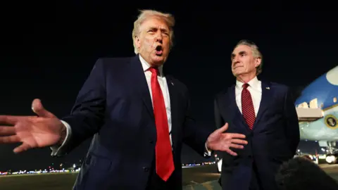 US President Donald Trump on the airport tarmac with Air Force One behind him. The president has his hands and mouth open as he speaks to a boom mic. Secretary of the Interior Doug Burgum stands to the president's left and watches him speak. 