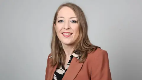 Dr Corrigan is smiling directly at the camera, she has shoulder length light brown hair. she is waring a tan coloured blazer an a black and white printed shirt.