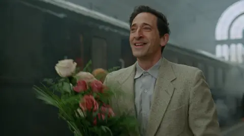 A24 Adrien Brody as László Tóth. He has short dark hair and is wearing a beige suit and grey shirt. He is holding a bunch of pink and white flowers and smiling as he walks along a train platform with a train behind him.