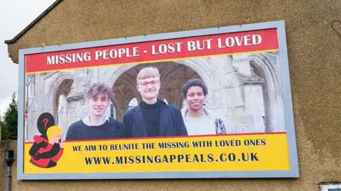 PA Media A billboard on the side of a building - large, with a picture showing Ben and two other people. There is a lot of red and yellow - it says "Missing People - Lost but Loved" and 'We aim to reunite the missing with loved ones'.