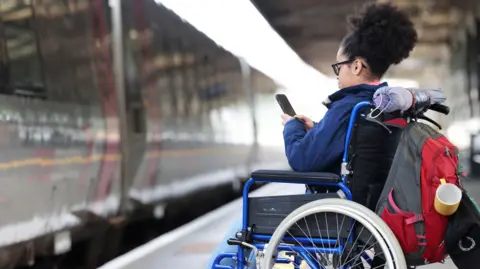 A woman in a wheelchair waiting to board a train, whilst using her phone.