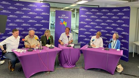 Picture of all the candidates sitting at the debate, from left to right: Andrew Pakes (Labour), Nick Sandford (Liberal Democrats), Nicola Day (Greens), Chris Mann (presenter), Paul Bristow (Conservative) and Sue Morris (Reform)