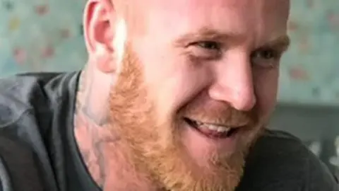 Family handout Lenny Scott, who has brown eyes, a shaven head, a ginger beard and a tattoo on his neck, smiles at the camera while wearing a grey t-shirt.