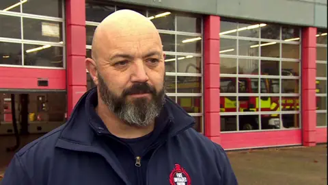 Mark Chapman has a dark brown beard and is wearing a blue jacket with a Fire Brigades Union while standing in front of a fire station. Fire engines can be seen through windows in the background.