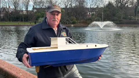 A man standing in front of a lake is wearing a navy blue jacket and a grey coloured cap. He is holding up his model boat, which has a blue hull and a white superstructure.