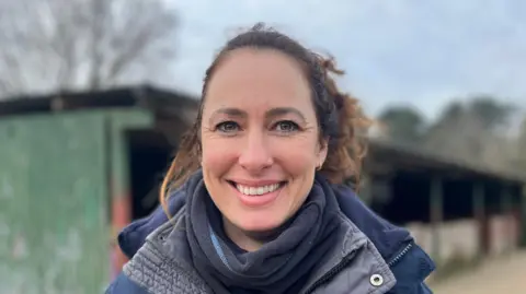 A woman with long brown hair tied up. She is stood in front of stables wearing a blue coat and scarf, while smiling at the camera.