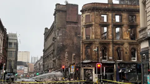 The remains of a building in Glasgow after the fire. A facade remains with nothing behind. The streets are covered in rubble and there is nobody in shot.