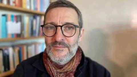 Alex looks directly at the camera with a slight smile. He has short greying hair, with a short grey and black beard and moustache. Alex wears thin round black wire frames glasses, a paisley printed red and yellow scarf tied around his neck and a black shirt jacket with the collar up. The background is softly focussed, but behind him there are rows of books on shelves.