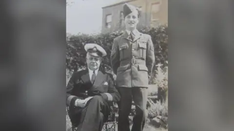 Brian Hughes A young man in his military uniform stands beside an old man in uniform who is sitting down. They are in a garden.