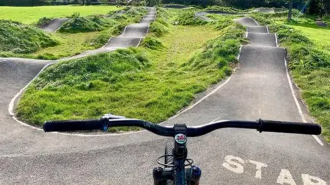 A bicycle is positioned at the start of a pump track with bumps, jumps and curves ahead, and the word Start can be seen near the handlebars.