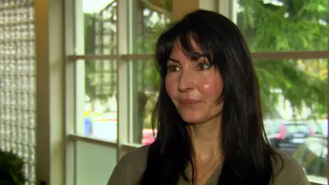 A woman with long dark hair, a green top and brown eyes looks off camera. 