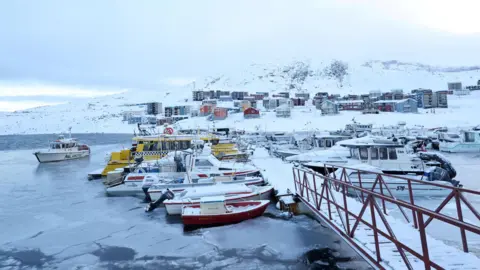 Reuters A view of a harbour in Greenland. Boats can be seen docked by a small pedestrian bridge and houses/buildings can be seen in the background. Everywhere is covered in snow and ice.