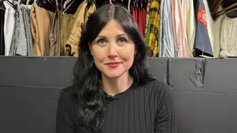 Jessica Bayley/BBC A woman with long dark hair and a black top wearing red lipstick. She is sitting against a black wall and there is a rail of clothes behind her. 