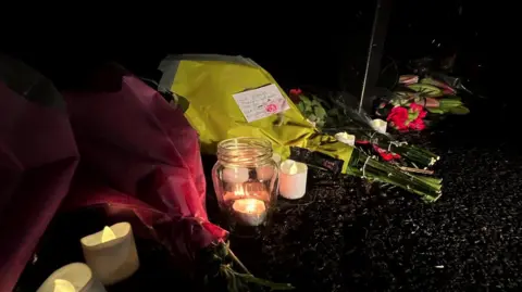 Bouquets of flowers have been placed alongside candles outside of the house. The flowers are wrapped in yellow and red paper.