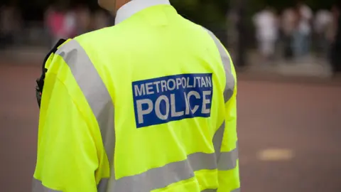 Stock image of a Metropolitan Police officer on patrol, in a yellow hi-viz, with the wording Metropolitan Police on the back. It is an anonymous picture, just with his back in shot. 