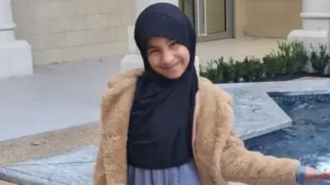 Inaayah wearing a brown furry jacket and a headscarf, smiles at the camera as she stands next to a water fountain in the shape of a star.