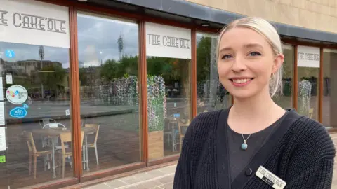 A young woman with scraped-back blond hair standing in front of a cafe's glass and wood-framed windows