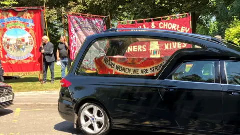 A black hearse with a coffin in the back covered by a coloured banner. In the background three other brightly coloured banners, synonymous with mineworkers, are held aloft
