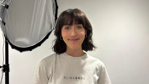 Jenna Buesnel está de pie en el interior, frente a una pared blanca lisa. Tiene el cabello oscuro hasta los hombros y lleva una camiseta blanca con el texto visible "SLINGSHOT FILMS". Una gran caja de luz circular para fotografía, colocada sobre un trípode, se encuentra a la izquierda de la imagen.