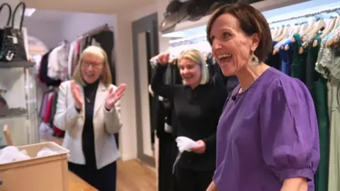Anne is in the foreground wearing a bright purple blouse and speaking with an animated smile to someone opposite. They are in a dress shop, and two women behind her are clapping and celebrating.