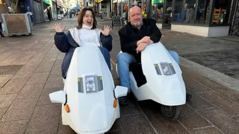 BBC/ITV Cymru Wales Ruth Jones and Steve Speirs on white scooters, Ruth has her hands in the air while Steve's hands are crossed and leaning on the scooter. They are parked in the middle of a street with shops on either side.