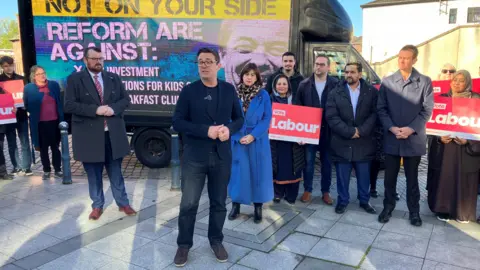 Andy Burnham in a dark suit and dark hair and dark rimmed glasses and Lucy Powell, with dark hair and a blue coat with Labour activists holding vote Labour red signs in Prestwich.