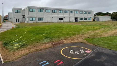 A grey two-storey building is in the background, behind a green field and play area.