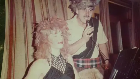 Barlow and Trevor Davies in fancy dress on stage. Barlow wears a long red wig and black woman's top, while Davies wears red tartan kilt, white shirt with a black sash, and a grey wig. Davies holds a microphone, with a stage curtain behind them both. 
