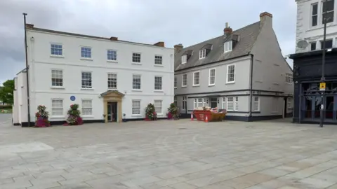 South Kesteven District Council A large white building with Georgian-style windows and Christmas trees along its frontage. Next to it is a large grey-coloured building with windows in the roof space. There is a large paved area in the foreground.