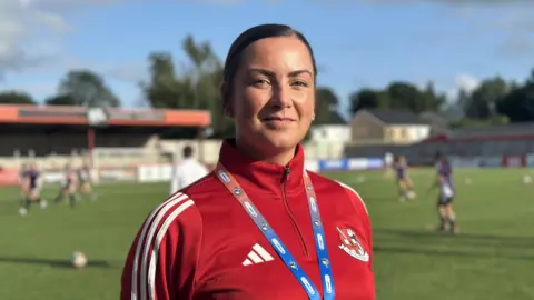 Clare McIlwaine has brunette hair which is tied back and she wears a red quarter zip sports jacket and a lanyard around her neck. She is standing on a football pitch and people can be seen playing football in the background.