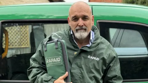 Alex Chivers stands in front of a Hackney cab wrapped in green Rapaid branding. He wears a green Rapaid rain coat and holds a Rapaid kit