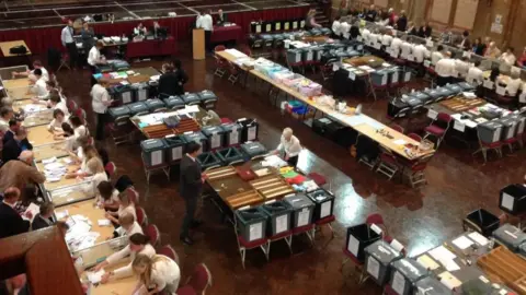 There are tables spaced out with ballot boxes and people counting votes at the tables. There are also candidates and agents watching over the tables.
