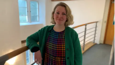 BBC Woman in green jumper stood in a hallway with right arm on a railing