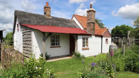 A small single storey cottage is located in a garden which has several flowers and trees. One side of the building is thatched and the other has red roofing tiles.
