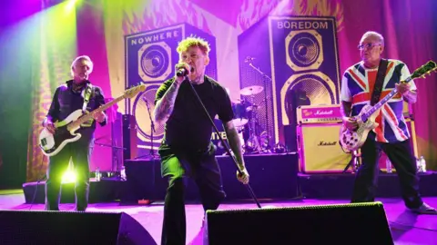 Getty Images A live band performs energetically on a brightly lit stage with colorful lights and amplifiers in the background. A central singer holds a microphone while two musicians on either side play guitars. Large speaker graphics behind them display the words “NOWHERE” and “BOREDOM,” adding a bold visual theme.