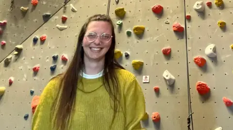 Alison Stephenson A woman is standing in front of an indoor climbing wall facility. She is wearing a baggy yellow jumper and has her hands down by her sides. She is smiling at the camera. 