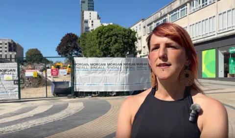 A woman with red hair in a black halter-neck top stands in a shopping centre. She is wearing large circular earrings and has a small microphone attached to her top. Behind her is a pedestrianised area and the area where the trees were felled. It is cordoned off and has hoardings around it which have the words Morgan Sindall printed on them.