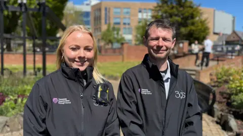A woman with blonde long hair and a man with short spicky brown hair smiling at camera. They are both wearing black jackets with logos of Causeway Coast and Glens Borough Council and 3GS. Sarah's jacket has a camera attached. There is a park in the background with people standing talking blurred in the background. 