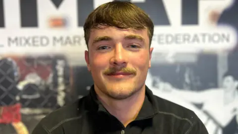 Ruairi O'Kane has brown hair and a moustache. He is smiling at the camera, wearing a black t-shirt.