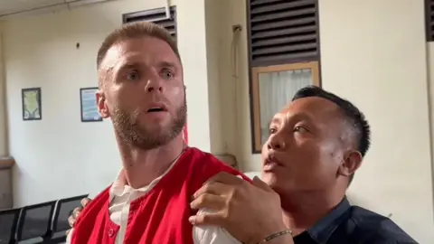 Thomas Parker being led into court. He has short brown hair and a beard and is wearing a red jacket and white shirt issued by the court. He is being held on the shoulder by an Indonesian man who works for the court.