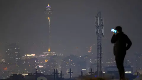 NurPhoto / Getty Images A man stood on a hill holds a phone to his ear, its screen can be seen glowing. Off in the distance are the lights of Tehran at night with a large communications tower, and a separate mobile phone mast in the distance. 