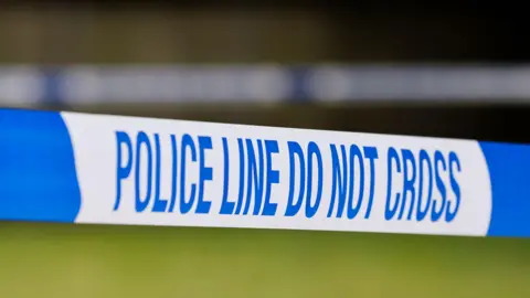 Taut blue-and-white police tape marked with the words: "Police line do not cross", against a blurred green background.