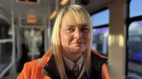 BBC Head and shoulders image of Holly Thompson on board a tram. She has long blonde hair and is wearing a high-vis orange jacket over a shirt and tie. She has a silver nose-ring.