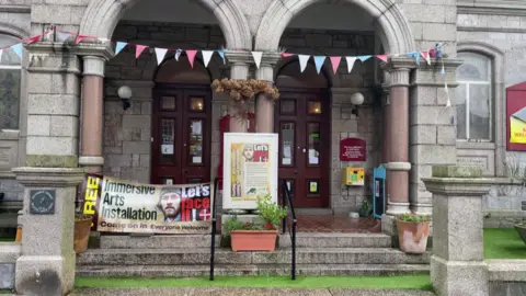 BBC The outside of a church building decorated with bunting and a sign for the immersive arts installation. Grass and steps in front of the two panelled door building.