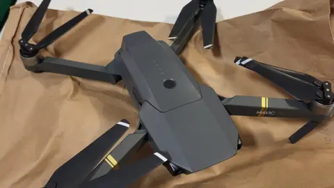 A black drone featuring four arms with propellers attached lies on a brown paper evidence bag on top of a table. 