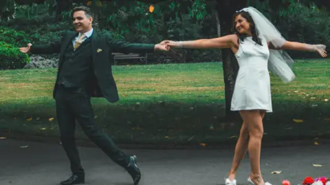 Felixine Hick Harmony and Seán smiling whilst dancing in the park. They are holding each others hand, stood in front of a tree and greenery. Harmony is looking at Seán smiling. 