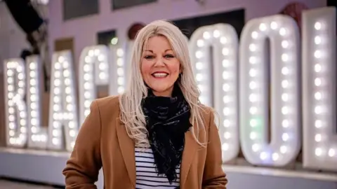 Blackpool Council Kate Shane standing in front of a white lightbulb-lit sign that says BLACKPOOL. She is smiling and has long straight blonde hair and is wearing a brown jacket over a striped top with a black scarf