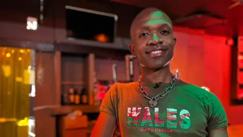 Bald man smiles at camera. He is wearing a green t-shirt with the Welsh flag on the word 'Wales, God's Heaven'