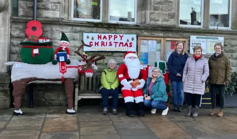 Suzanne Excell A knitted reindeer is pictured next to a bench on which sits one of the knitters and a smaller, knitted Santa, while the other knitters crouch and stand nearby