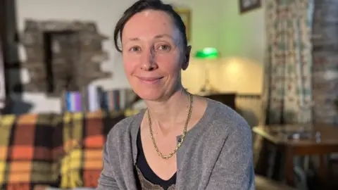 Dr Hart sitting on her sofa wearing a grey cardigan and gold necklace